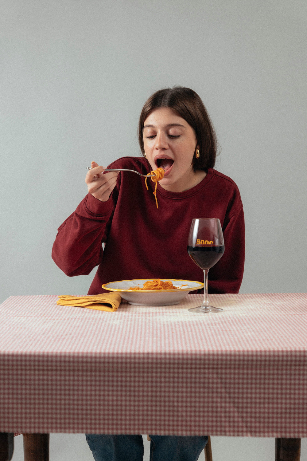 Une personne mange des spaghetti à une table à carreaux, en sweat bordeaux.